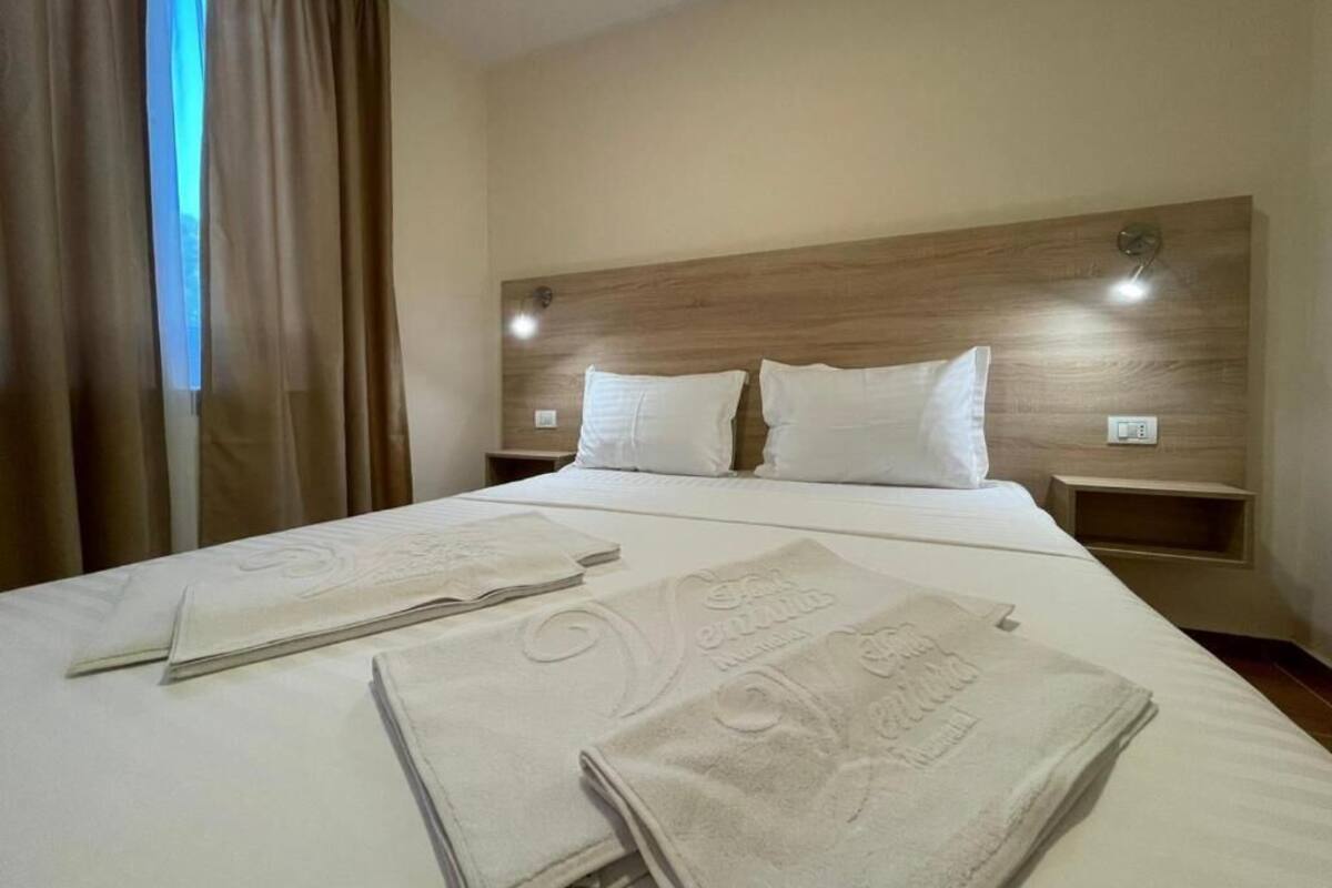 Ein Hotelzimmer mit einem großen Bett, zwei Kissen, einem Kopfteil, Nachttischen und einem fenster mit Vorhängen.