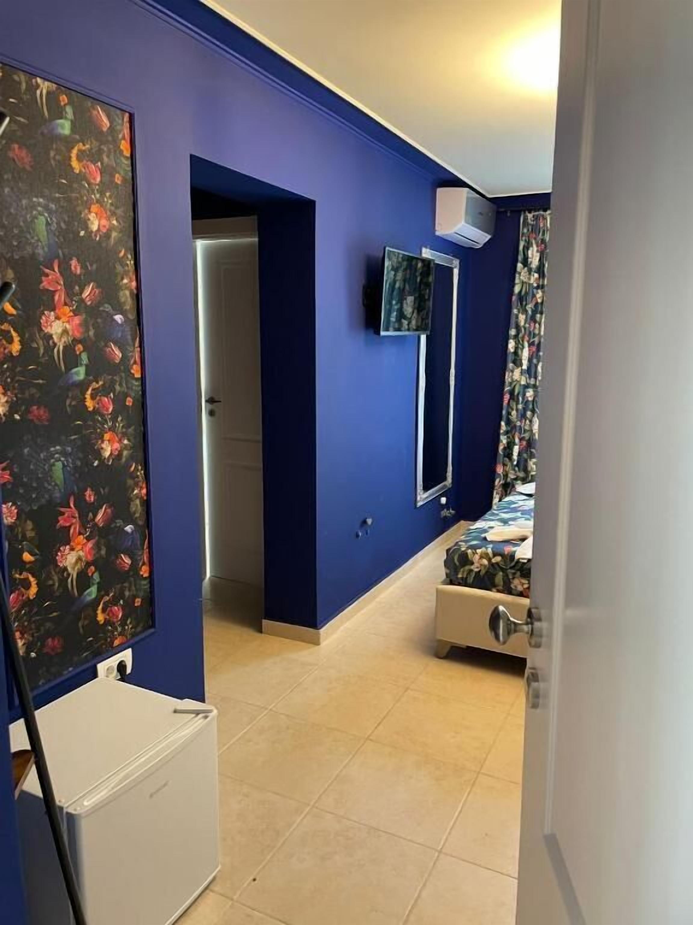 Una habitación con paredes azules, una cama con ropa de cama floral, un refrigerador blanco y un televisor montado en la pared.