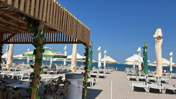 A beachside area with rows of white lounge chairs and umbrellas, a wooden pergola, and a trash bin labeled 'Rose Veritas.' - Hotel Olt by Ventura (Mamaia)