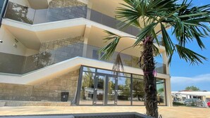 A modern building with a glass entrance, a palm tree, and a black outdoor seating area. - Hotel Olt by Ventura (Mamaia)