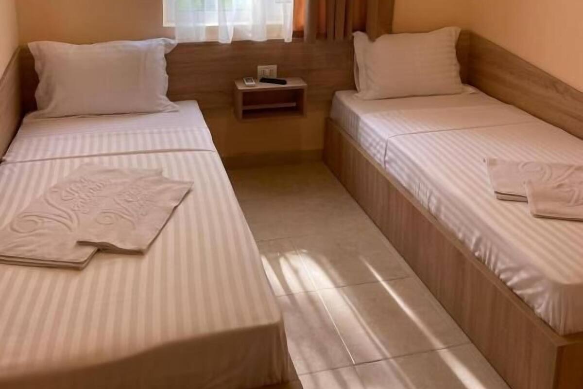 Ein Zimmer mit zwei Einzelbetten, einem kleinen Nachttisch mit Lampe und einem fenster mit Vorhängen.