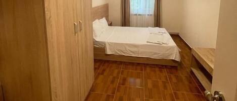 Habitación de hotel con armario de madera, cama con colcha blanca, televisor de pantalla plana montado en la pared y ventana con cortinas.