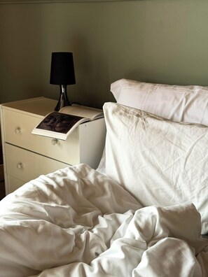 Una cama con ropa de cama blanca, una mesita de noche con una lámpara y un libro.