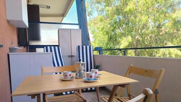 Ein Balkon mit Holztisch und -stühlen, einer kleinen Küchenzeile und Blick auf Bäume.