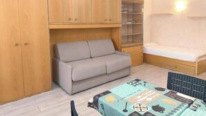 Ein kleiner Raum mit einer Schlafcouch, einem Bett, einem Schreibtisch und Holzschränken.