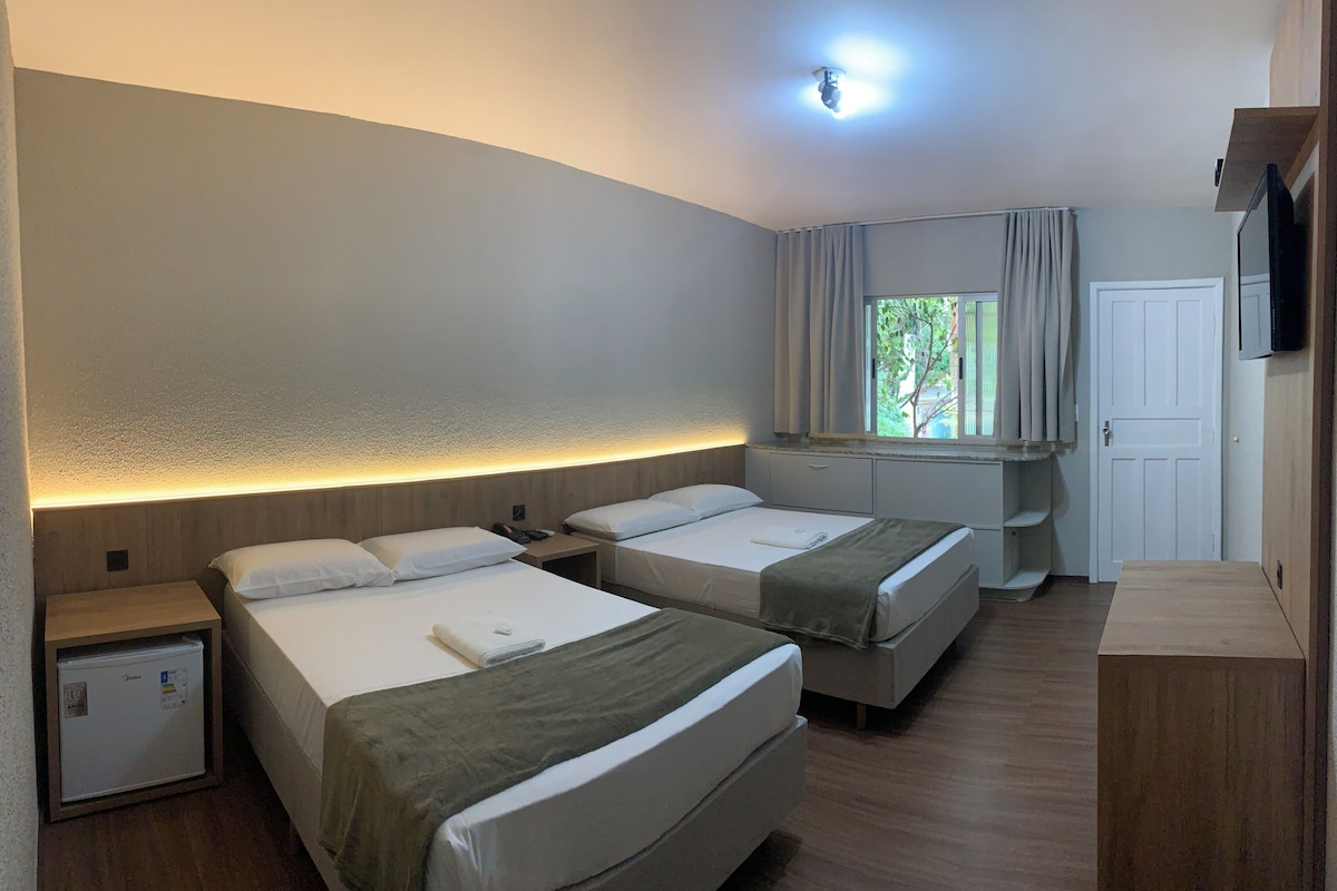 Quarto de hotel com duas camas, uma escrivaninha pequena, televisão e uma janela com cortinas.
