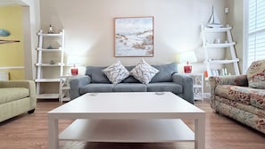 Ein Wohnzimmer mit grauem Sofa, zwei gemusterten Kissen, einem weißen Couchtisch und einem Bild einer Strandansicht an der Wand.