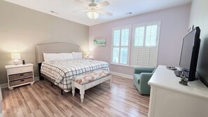 Ein Schlafzimmer mit Bett, Nachttischen, einem Schreibtisch, einem Stuhl, einem Deckenventilator und einem Fenster mit Jalousien.