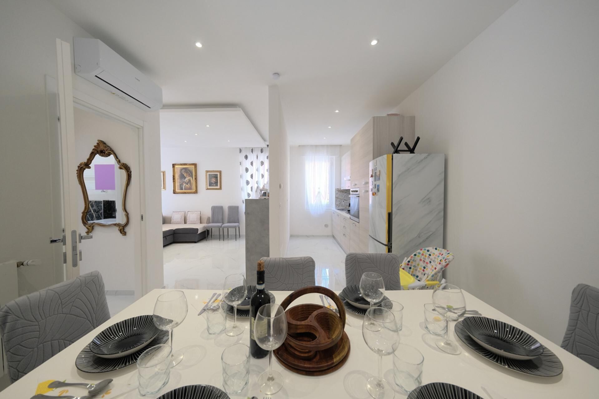 Uma sala de jantar moderna com uma mesa branca posta para seis, cozinha ao fundo e sala de estar com sofá e poltronas.