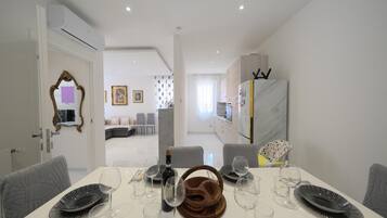 Uma sala de jantar moderna com uma mesa branca posta para seis, cozinha ao fundo e sala de estar com sofá e poltronas.