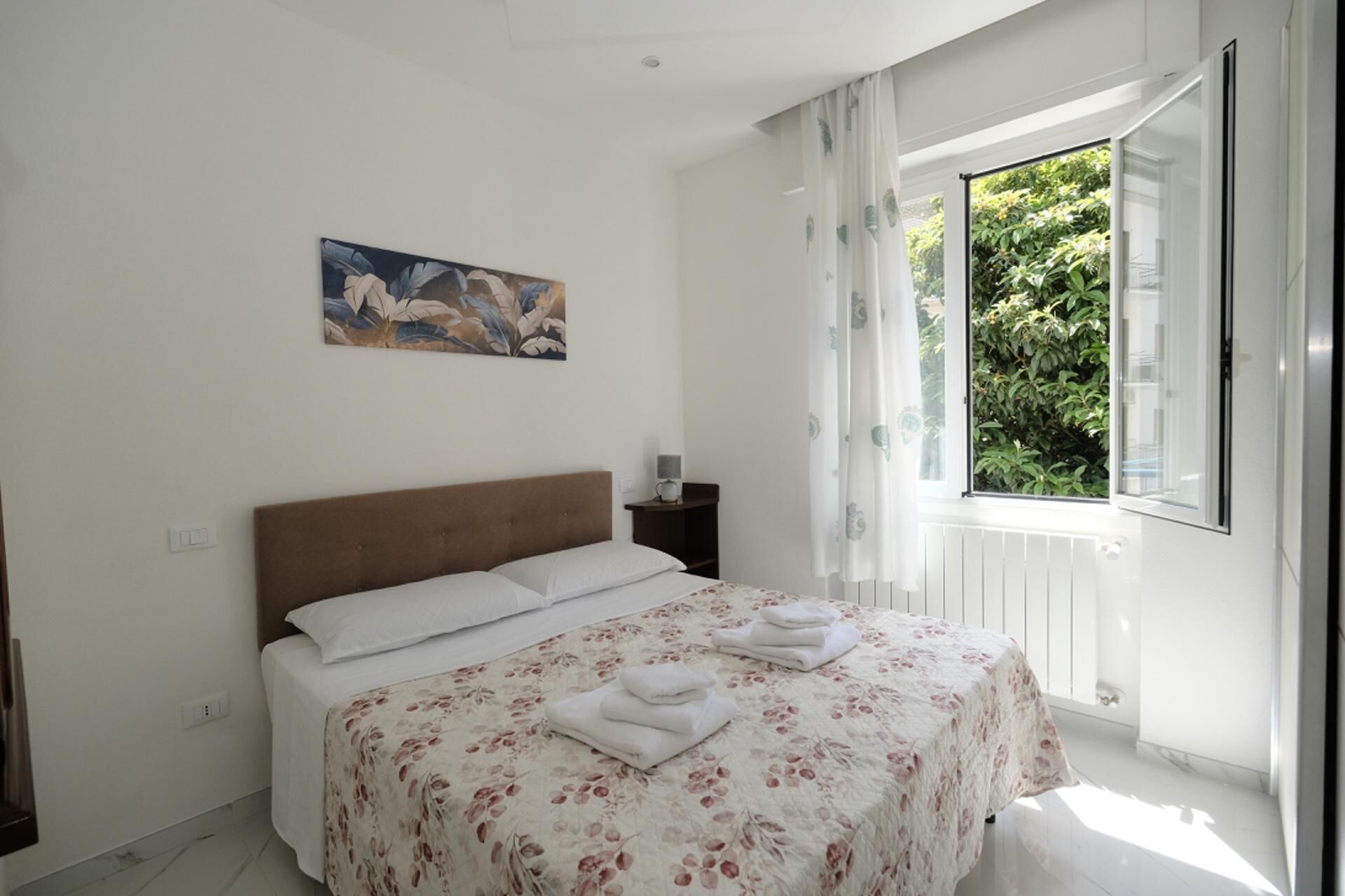 Un dormitorio con cama, mesita de noche, un cuadro y una ventana con cortinas.