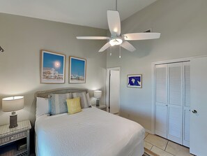 A bedroom with a bed, a ceiling fan, two framed pictures, a nightstand with a lamp, and a door with louvered shutters. - The Landing Gulf Shores #104 | Beach + Lagoon Access + Pool (Gulf Shores)