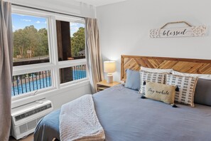 Ein Schlafzimmer mit großem Fenster, einem Bett mit Kopfteil, einem Nachttisch mit Lampe und einer Wanddekoration.
