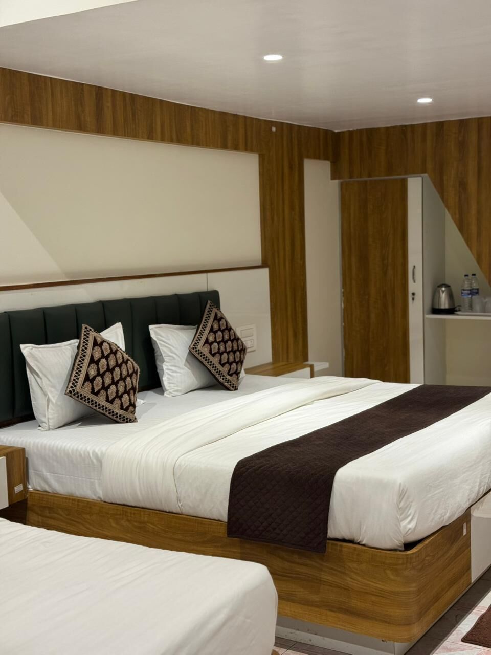A modern hotel room with a large bed, a wooden headboard, a wardrobe, and a small table with a kettle.