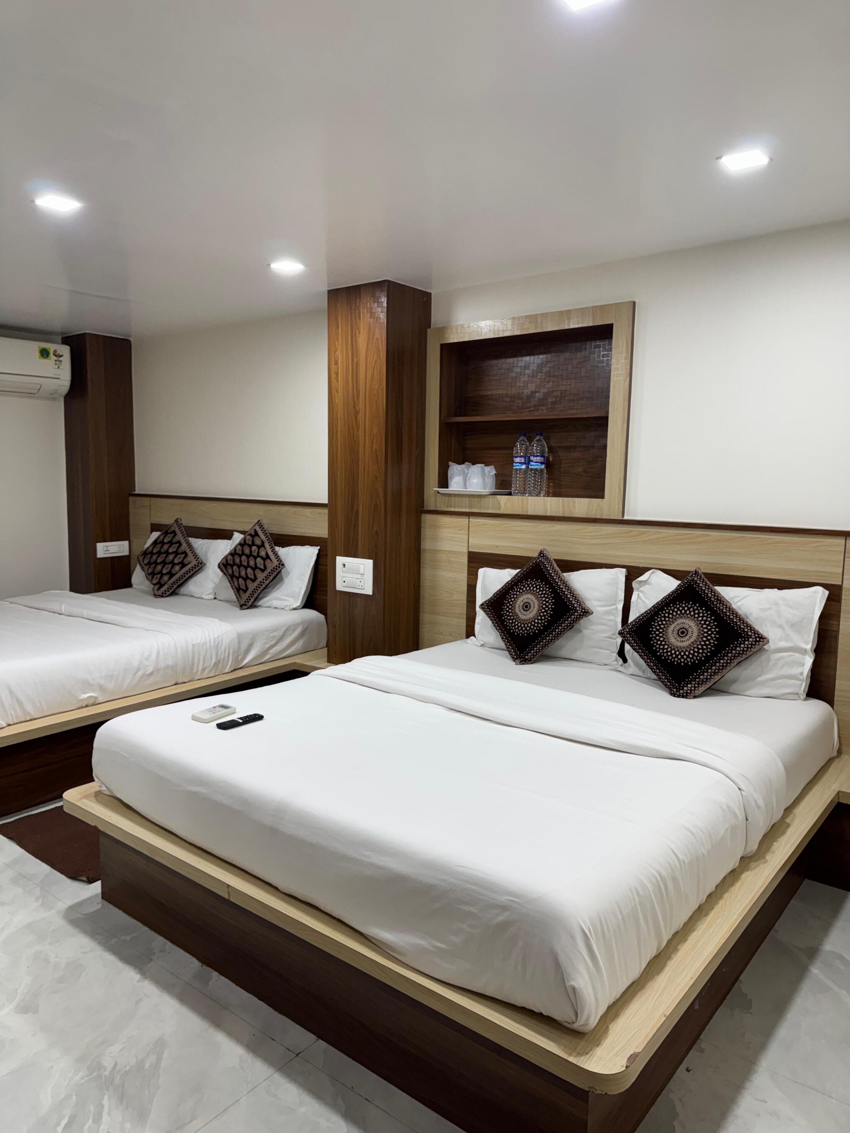 Une chambre d’hôtel avec deux lits, une tête de lit en bois, une étagère avec des bouteilles d’eau et un climatiseur fixé au mur.
