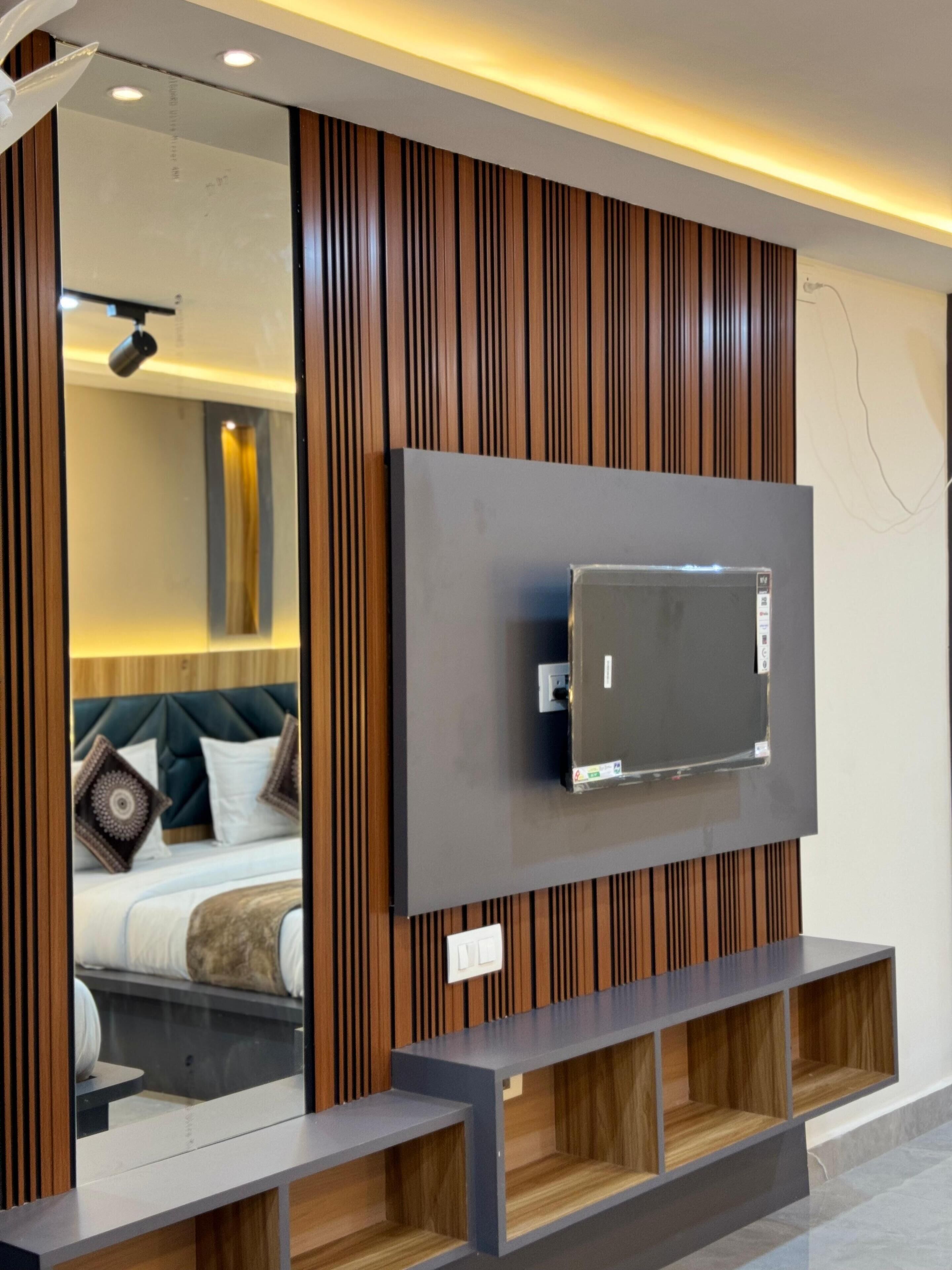 Una habitación de hotel moderna con un televisor de pantalla plana grande montado en una pared de paneles de madera, una cama con cabecera y un estante empotrado debajo del televisor.