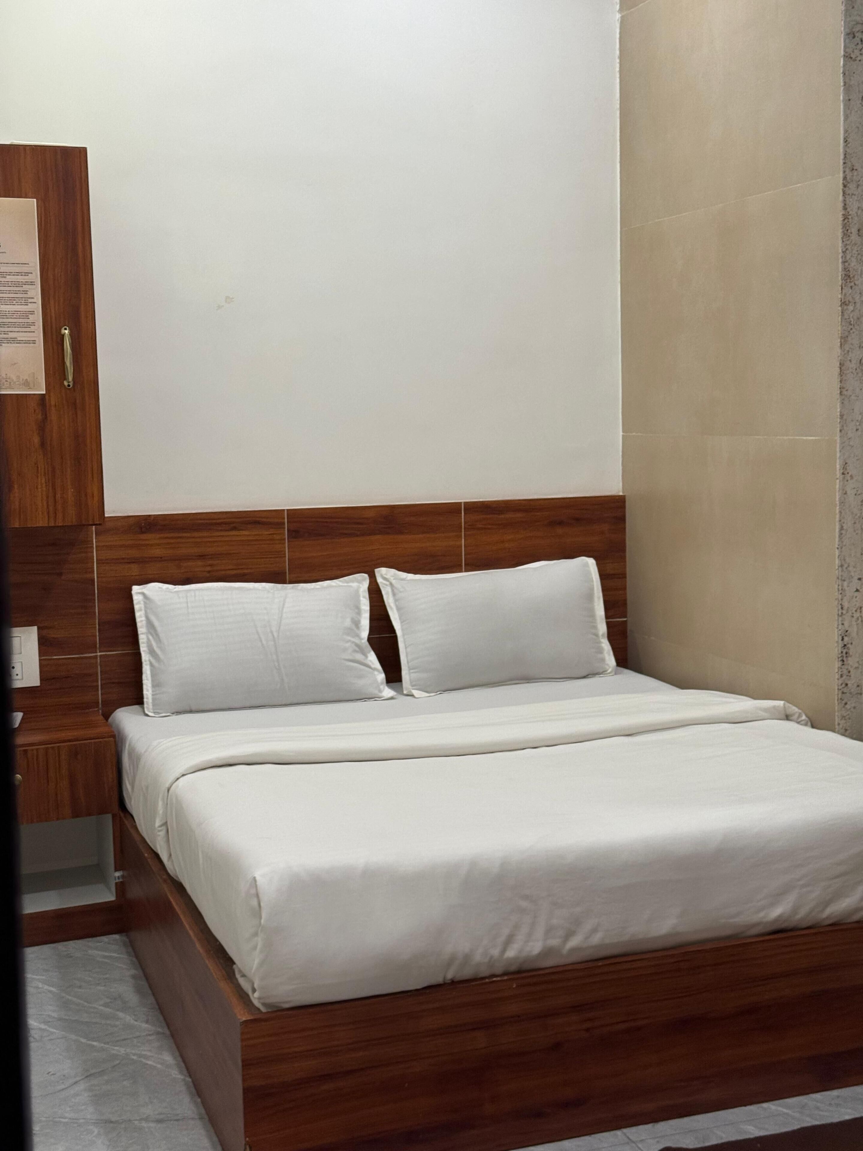 A neatly made bed with a wooden headboard, a small bedside table, and a wall-mounted light switch.