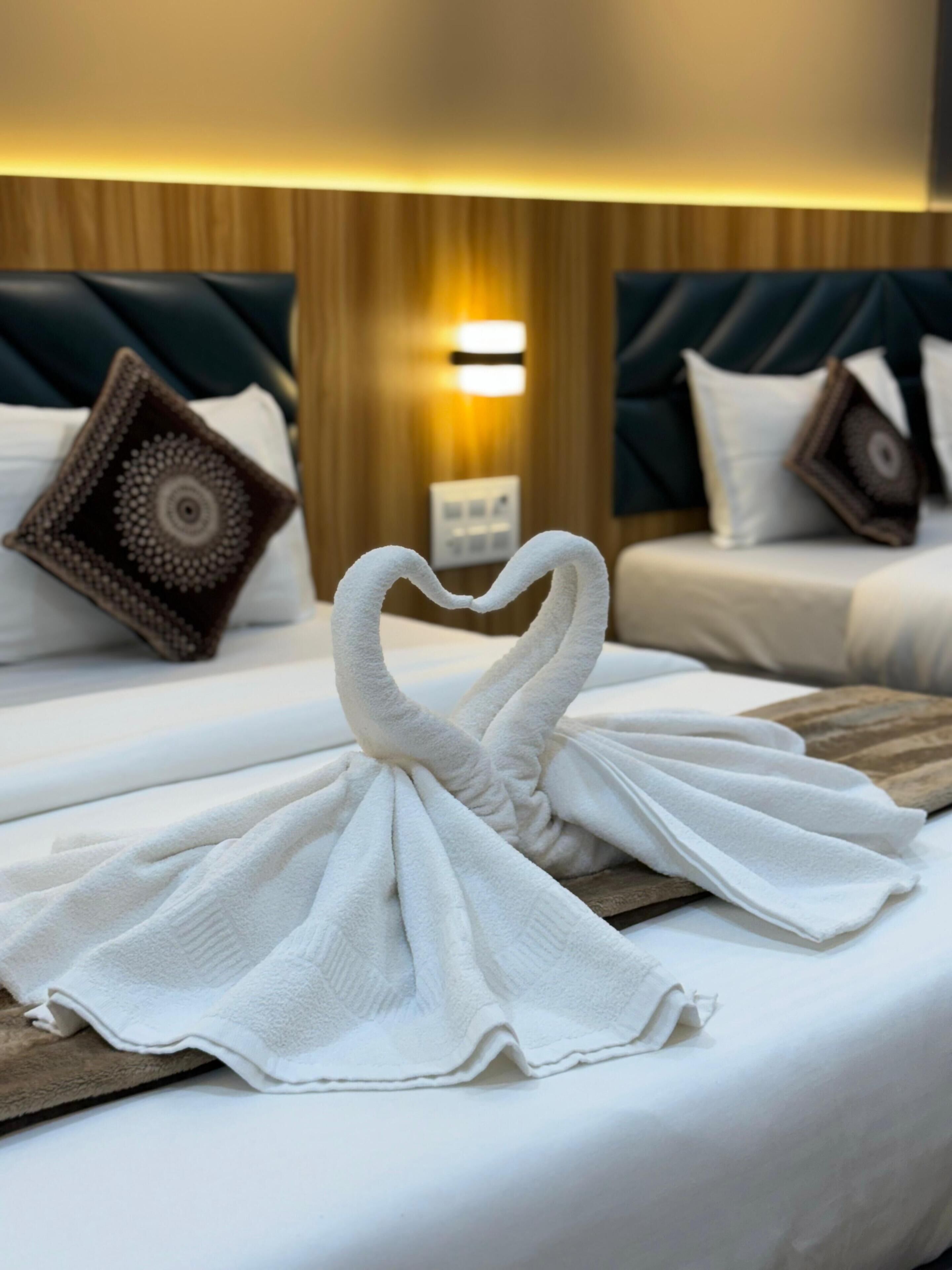 Une chambre d’hôtel avec un lit agrémenté d’une décoration en forme de cœur réalisée avec des serviettes, une tête de lit en bois et une lampe fixée au mur.