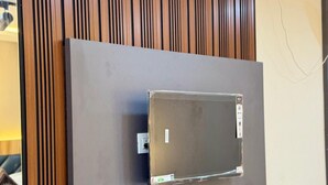 A flat-screen TV mounted on a wall with a wooden panel design, a power outlet, and a shelf below. - Yatri Stay inn (Mumbai)