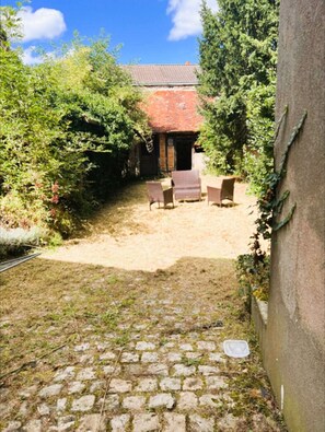 Une cour bordée d’un mur de briques, un petit bâtiment avec un toit rouge et un espace aménagé avec des chaises.