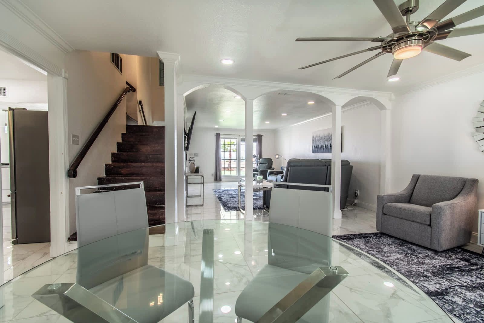 A modern living room with a glass-top dining table, a gray sofa, and a ceiling fan.