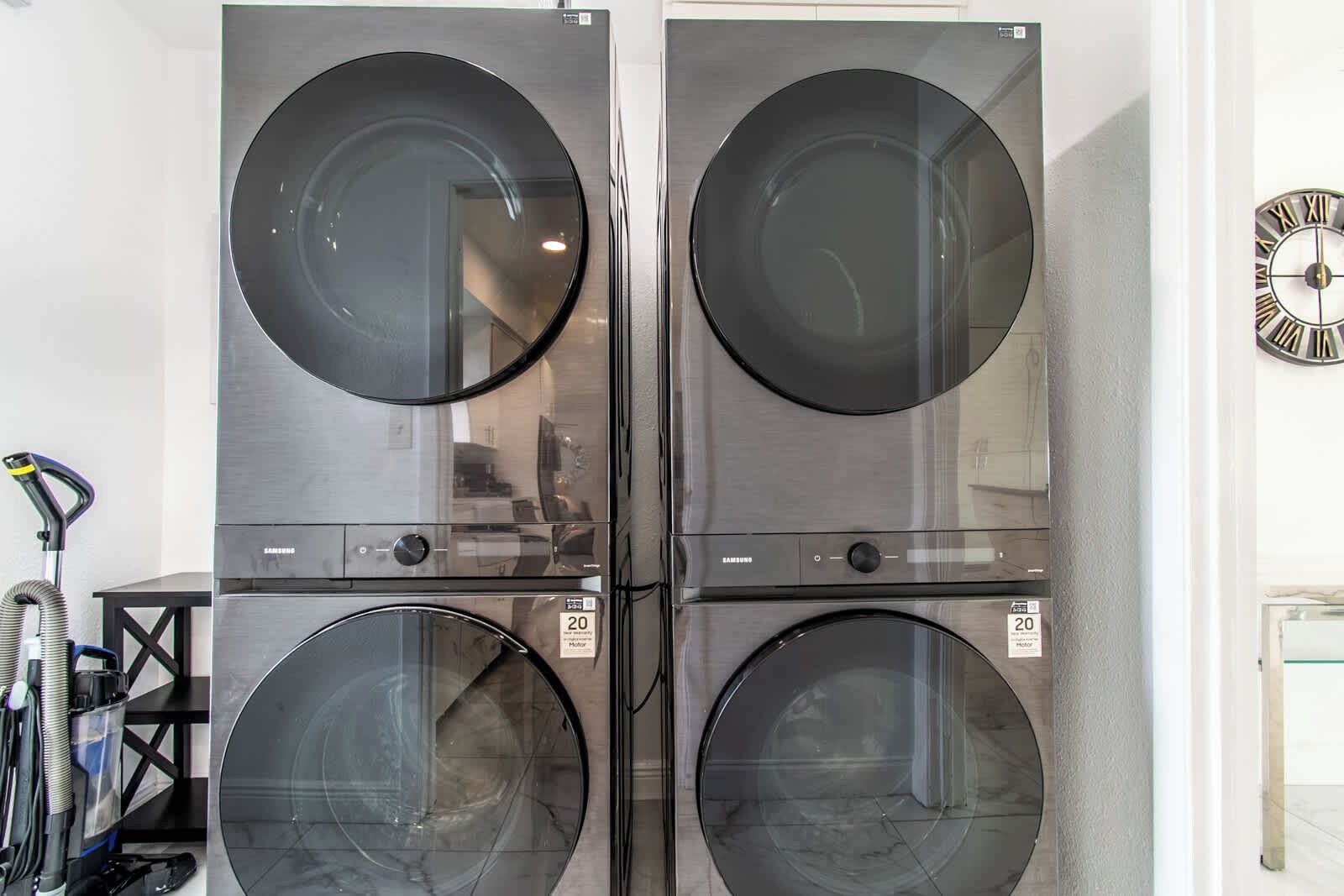 Quattro lavatrici a carica frontale in acciaio inox con superficie riflettente, esposte in una vetrina, con un aspirapolvere e un orologio a parete.