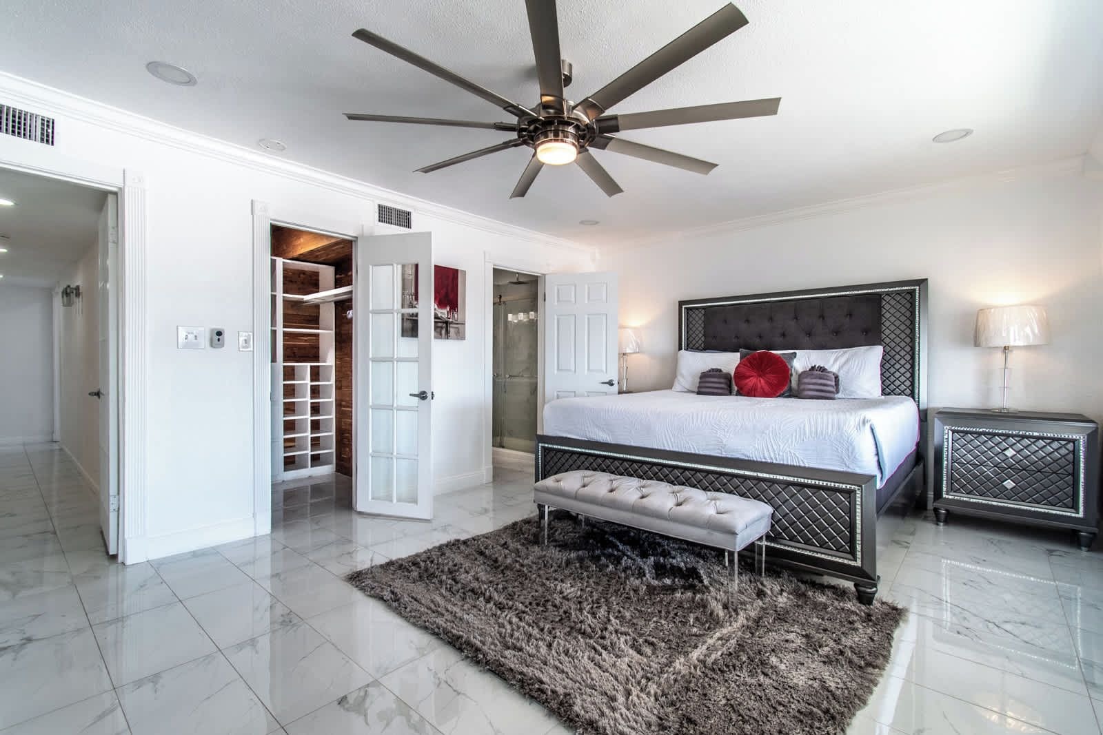Un dormitorio con cama, ventilador de techo, banco, mesita de noche y vestidor.