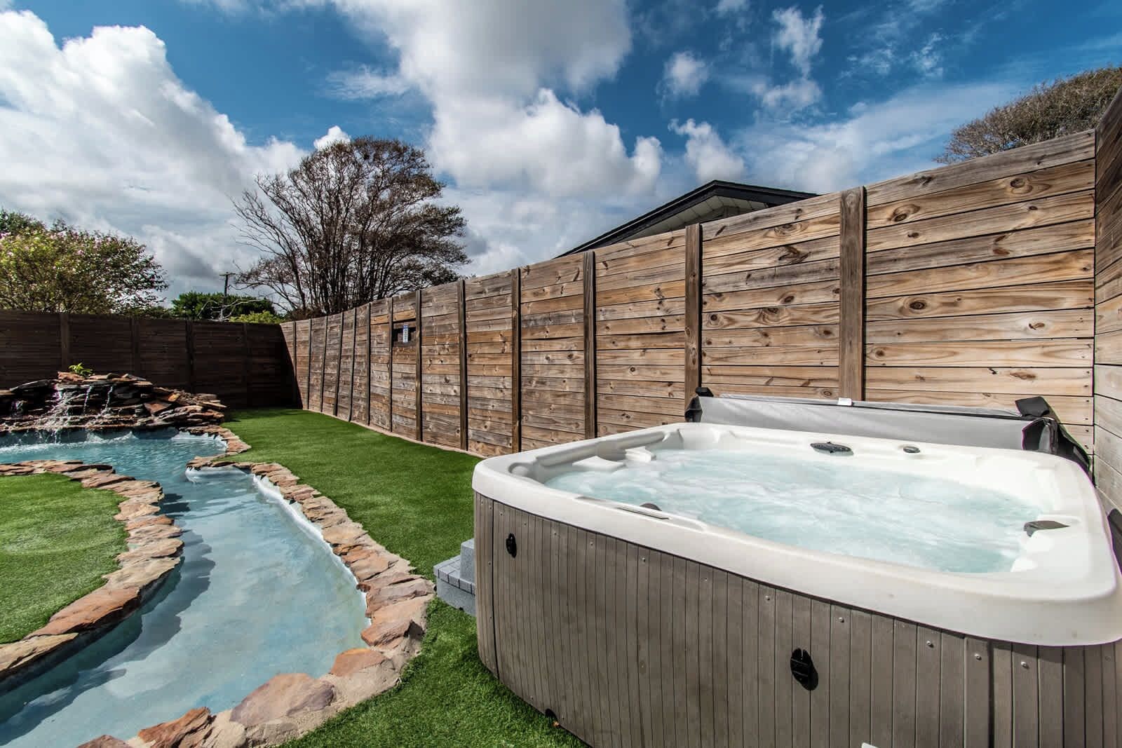Een achtertuin met een hottub, een kleine vijver en een houten omheining.