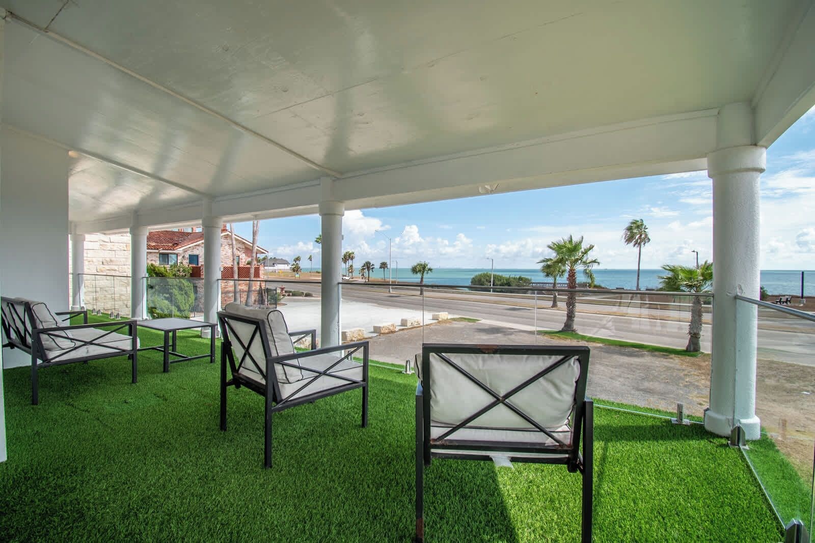 A covered outdoor seating area with artificial grass, overlooking a road and a body of water.
