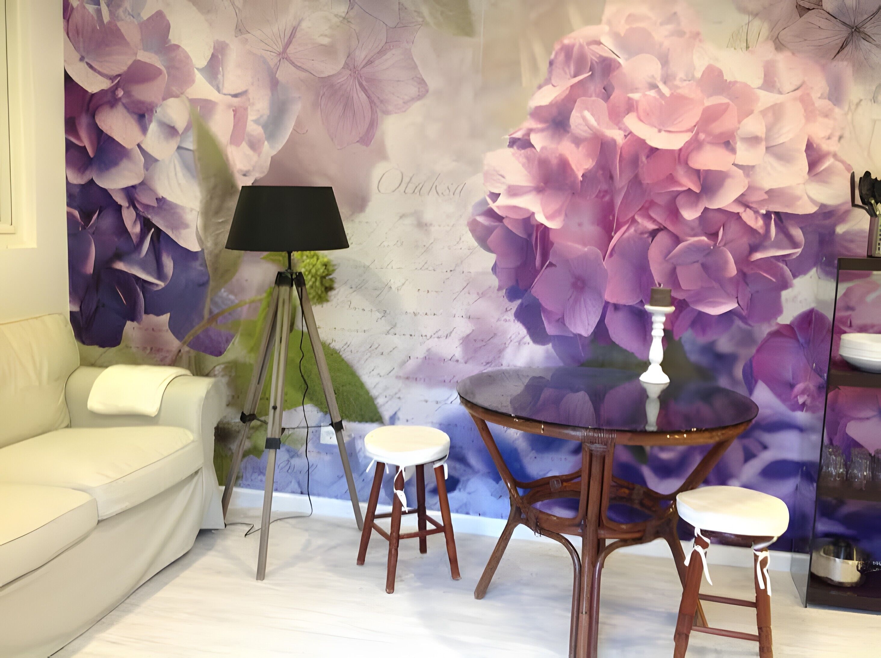 Et værelse med en hvid sofa, en tripodlampe, et rundt bord med barstole og en væg med et blomstermural.