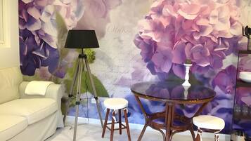 Et værelse med en hvid sofa, en tripodlampe, et rundt bord med barstole og en væg med et blomstermural.