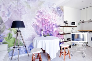 Ein Zimmer mit einem runden Tisch, zwei Hocker, einer Lampe, einem großen Blumenwandbild, einer Küchenzeile mit einer Mikrowelle, einer Waschmaschine und einem Fernseher auf einem Schrank.