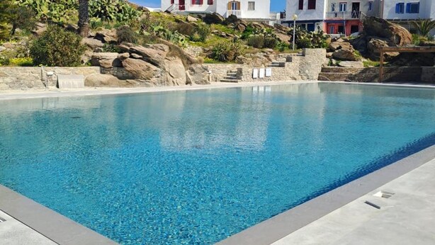 Une piscine à l’eau d’un bleu limpide, entourée d’une terrasse en béton. Au fond, on aperçoit des bâtiments blancs aux fenêtres rouges et bleues, qui se dressent sur une colline parsemée de verdure et de rochers.