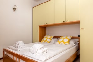 Ein Einzelbett mit weißer Bettwäsche und gelben Kopfkissen in Punkten, ein hölzernes Kopfstück und ein heller Wandschrank mit zwei Türen.
