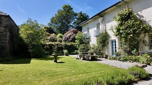 Um jardim bem cuidado com uma fonte de pedra, uma mesa de piquenique e um edifício com fachada branca e persianas azuis.