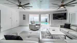 A spacious living room with a large TV, ceiling fans, and a view of the outdoors. - Ocean View + Poolside Opulence Tennis (Corpus Christi)