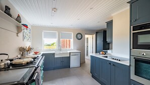 A modern kitchen with a stove, oven, and dishwasher, a window with a view of greenery, and a clock on the wall. - Shammer Lodge, North Creake, Norfolk (North Creake)
