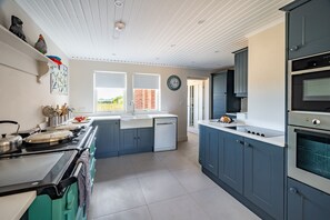 A modern kitchen with a stove, oven, and dishwasher, a window with a view of greenery, and a clock on the wall. - Shammer Lodge, North Creake, Norfolk (North Creake)