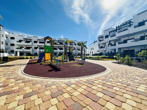 Ein Spielplatz mit bunten Spielgeräten, umgeben von einer gepflasterten Fläche mit gemusterten Fliesen.