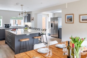 A modern kitchen with a central island, bar stools, and a dining table set for two. - Manor Farm Lodge, Brancaster, Norfolk (Brancaster)