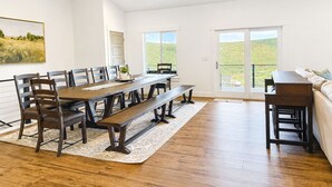 Un espace repas comprenant une longue table, des bancs et des chaises, un canapé, et une vue sur l’extérieur grâce à une porte vitrée.