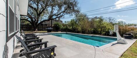 A swimming pool with lounge chairs, a basketball hoop, and a building in the background.