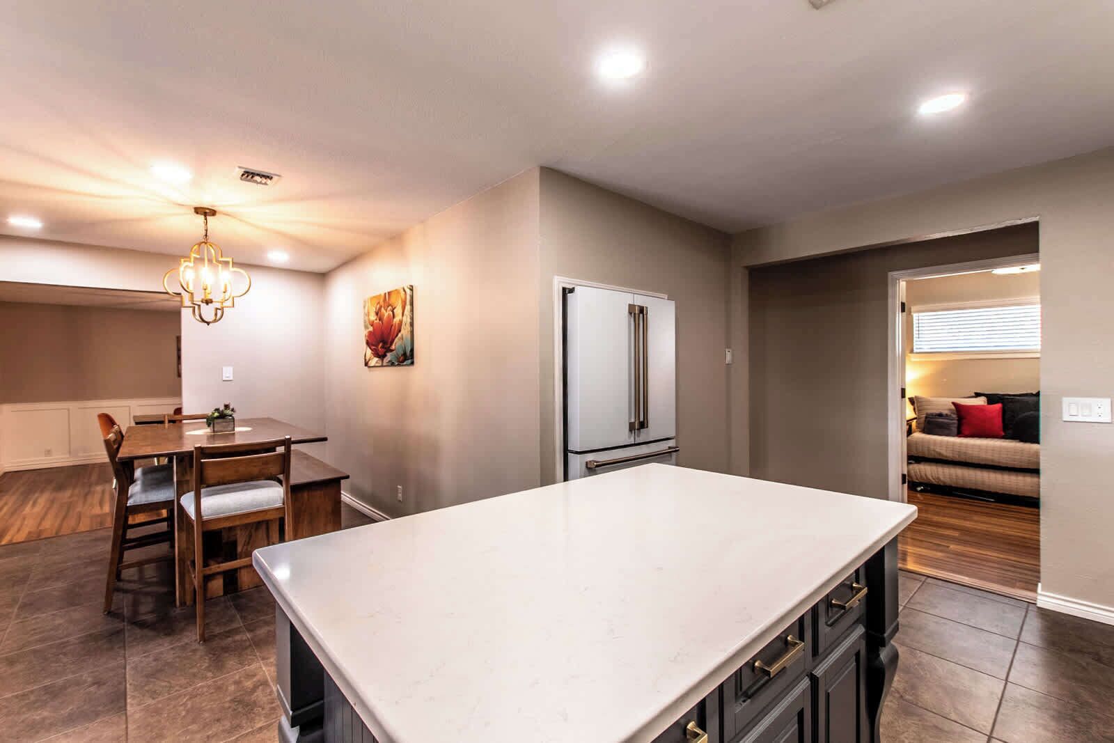 A modern kitchen with a central island, a dining area with wooden chairs, a refrigerator, and a painting on the wall.