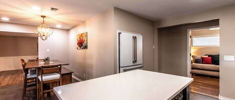 A modern kitchen with a central island, a dining area with wooden chairs, a refrigerator, and a painting on the wall.