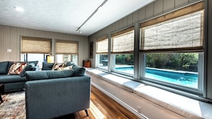 Een moderne woonkamer met een modulaire bank, grote ramen met zonwering en uitzicht op een zwembad.