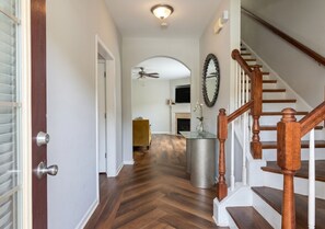 Un couloir avec un plancher en bois, une porte en verre, un ventilateur de plafond, un escalier avec des rampes en bois et un miroir rond accroché au mur.