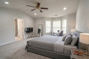 Une chambre à coucher comprenant un lit, un téléviseur, un ventilateur de plafond, une fenêtre avec des stores et une porte donnant sur une autre pièce.