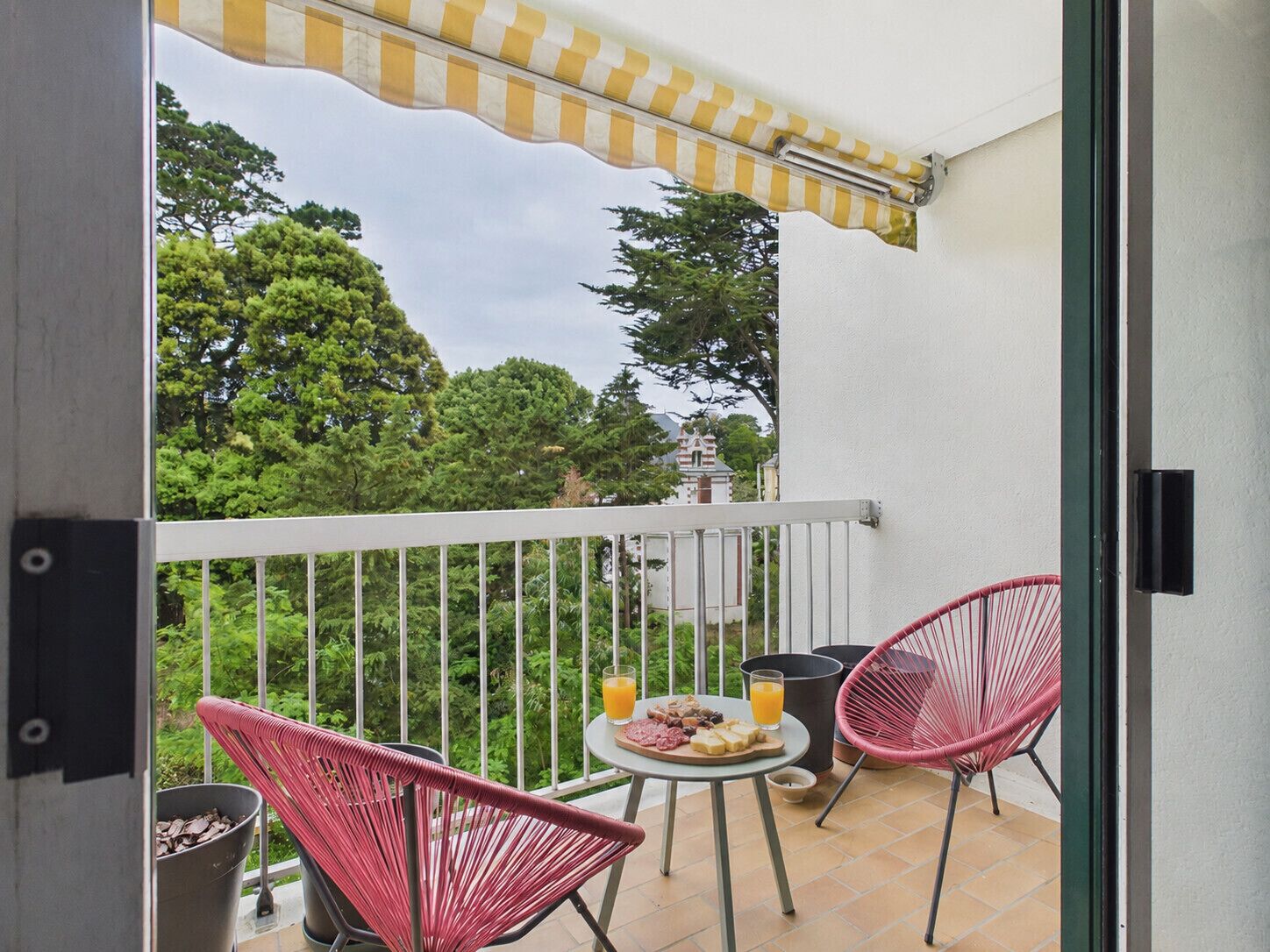 A balcony with a checkered awning, two wicker chairs, a small round table with food and drinks, and a view of trees and a building in the background.