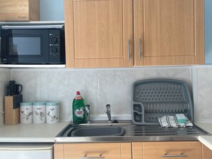 A kitchen with wooden cabinets, a microwave, a sink with a dish drying rack, and containers labeled tea, sugar, and coffee. - Apartment 6 (Torquay)