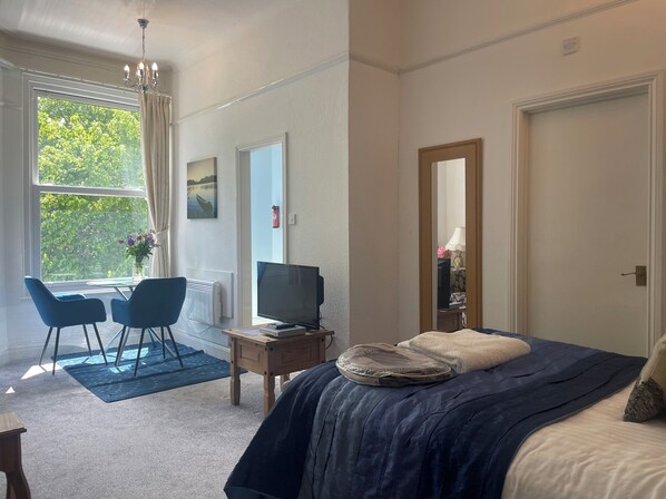 A hotel room with a bed, a desk with a chair, a television, and a large window with curtains. - Apartment 6 (Torquay)